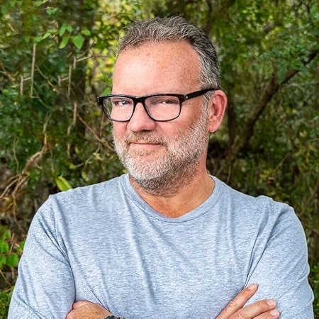 Portrait of a man with glasses and a grey beard in front of green foliage.